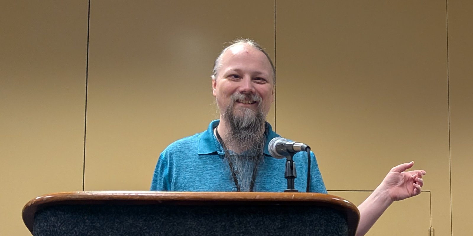 Jacob, wearing a teal polo, stands at a microphone behind a wooden podium. He is gesturing with one hand and smiling.